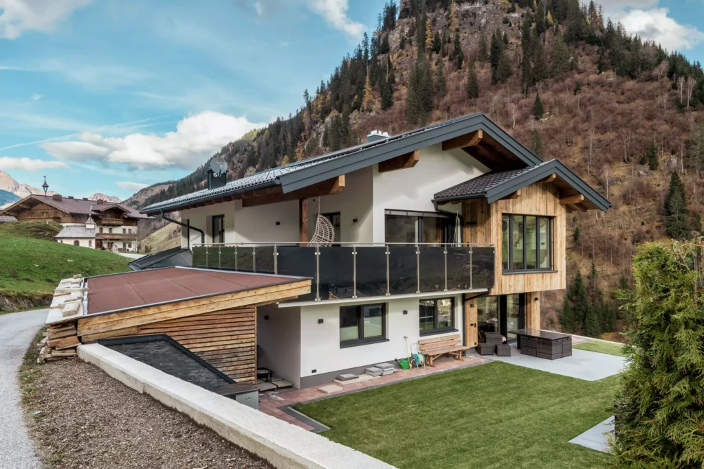 deutinger-projektentwicklung-baumanagement-generalunternehmen-immobilien-salzburg-MFH-Grossarl-Gfrerer-Schwarb-1 Modernes Mehrfamilienhaus mit Holz- und Putzfassade vor bewaldetem Berg in Großarl, Salzburg, mit Terrasse und Gartenbereich