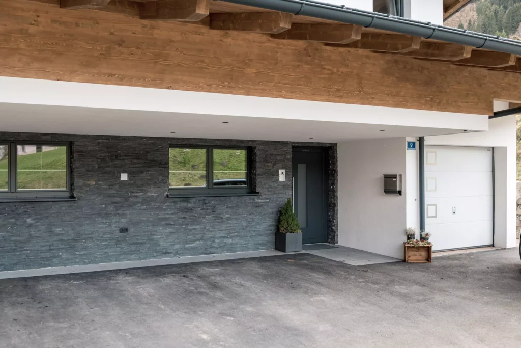 deutinger-projektentwicklung-baumanagement-generalunternehmen-immobilien-salzburg-MFH-Grossarl-Gfrerer-Schwarb-11 Eingangsbereich eines modernen Mehrfamilienhauses mit grauer Steinwand, dunkler Tür, zwei Fenstern und weißer Garagentür in