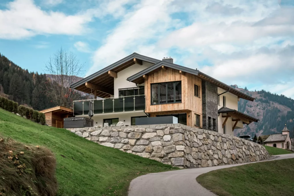 deutinger-projektentwicklung-baumanagement-generalunternehmen-immobilien-salzburg-MFH-Grossarl-Gfrerer-Schwarb-12 Modernes Mehrfamilienhaus mit Holzfassade auf steinerner Hangmauer in Großarl, umgeben von Bergen und grüner Landschaft