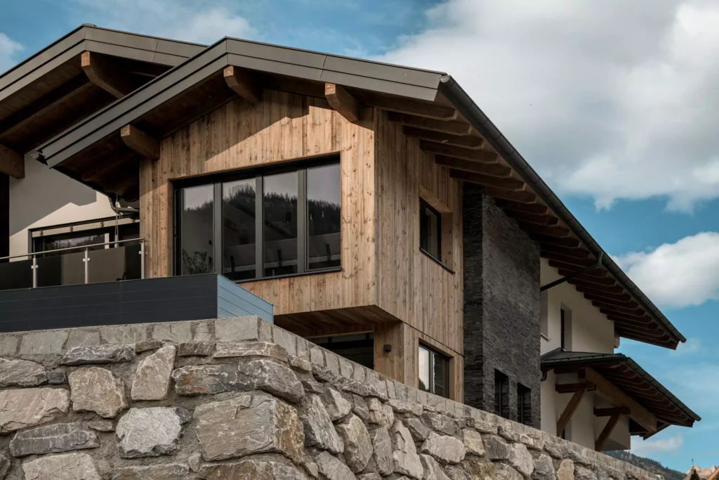deutinger-projektentwicklung-baumanagement-generalunternehmen-immobilien-salzburg-MFH-Grossarl-Gfrerer-Schwarb-3 Modernes Mehrfamilienhaus mit Holzfassade und Natursteinmauer im Berggebiet bei Großarl unter blauem Himmel