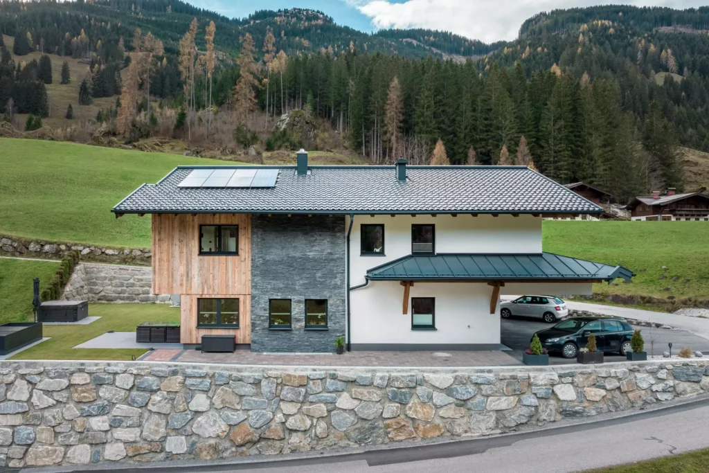 deutinger-projektentwicklung-baumanagement-generalunternehmen-immobilien-salzburg-MFH-Grossarl-Gfrerer-Schwarb-5 Modernes Mehrfamilienhaus mit Holz- und Steinfassade, Solarpaneelen auf dem Dach, umgeben von Bergen und Wald in Großarl