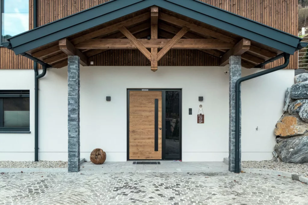deutinger-projektentwicklung-baumanagement-generalunternehmen-immobilien-salzburg-MFH-Grossarl-Wagnermeyer-11 Eingangsbereich eines modernen Mehrfamilienhauses mit Holzverkleidung, Steinsäulen und gepflastertem Vorplatz in Großarl.