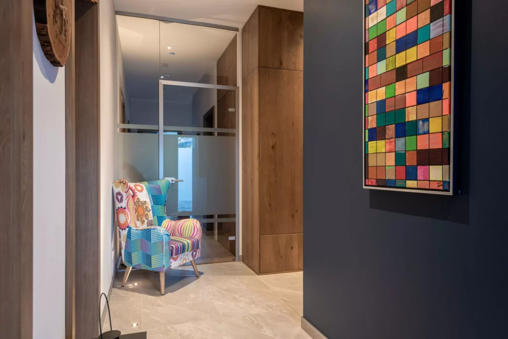 deutinger-projektentwicklung-baumanagement-generalunternehmen-immobilien-salzburg-MFH-Grossarl-Wagnermeyer-12 Moderner Flur mit buntem Sessel, Holzeinbauschrank, Glastür und farbenfrohem Wandbild in einem Wohngebäude