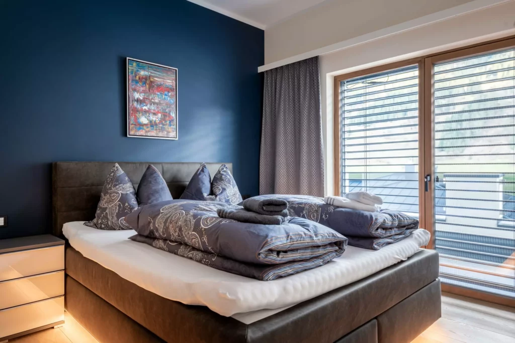deutinger-projektentwicklung-baumanagement-generalunternehmen-immobilien-salzburg-MFH-Grossarl-Wagnermeyer-14 Modernes Schlafzimmer mit Doppelbett, gemusterten Bettdecken, Nachttisch und großem Fenster mit Jalousien und Vorhang