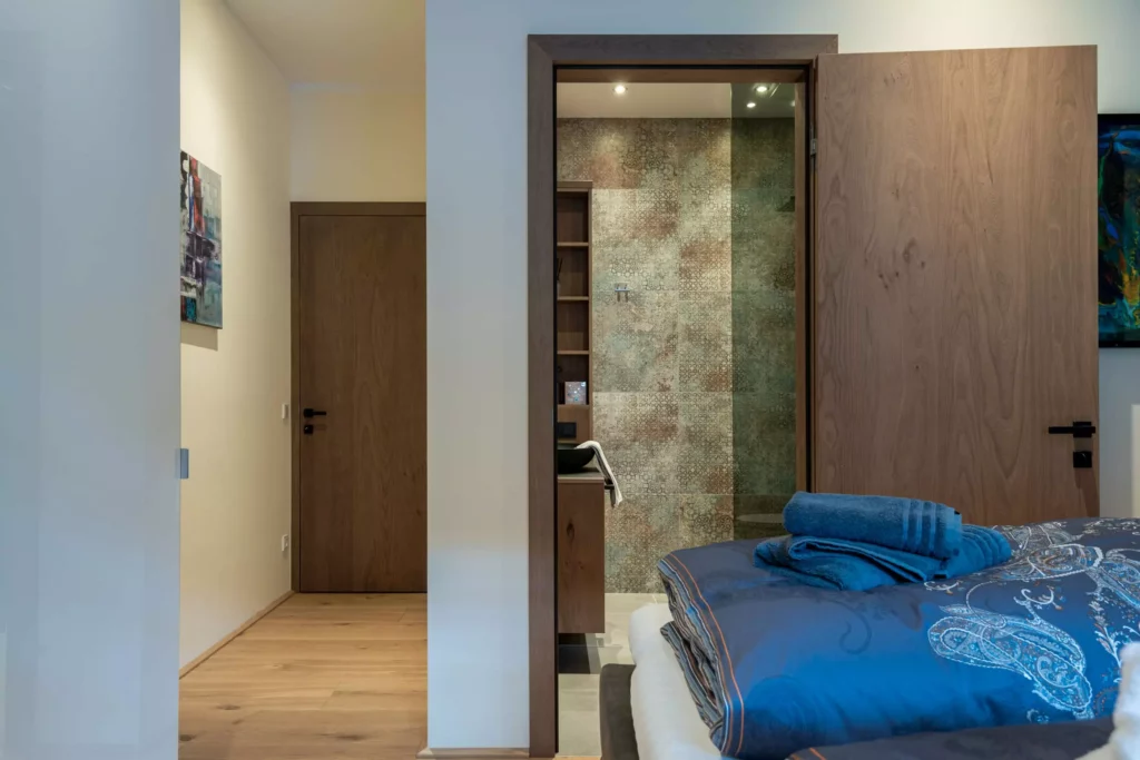 deutinger-projektentwicklung-baumanagement-generalunternehmen-immobilien-salzburg-MFH-Grossarl-Wagnermeyer-15 Blick ins Schlafzimmer mit blau gemustertem Bett und offenem Holztürrahmen zur Dusche mit gemusterten Fliesen im Bad