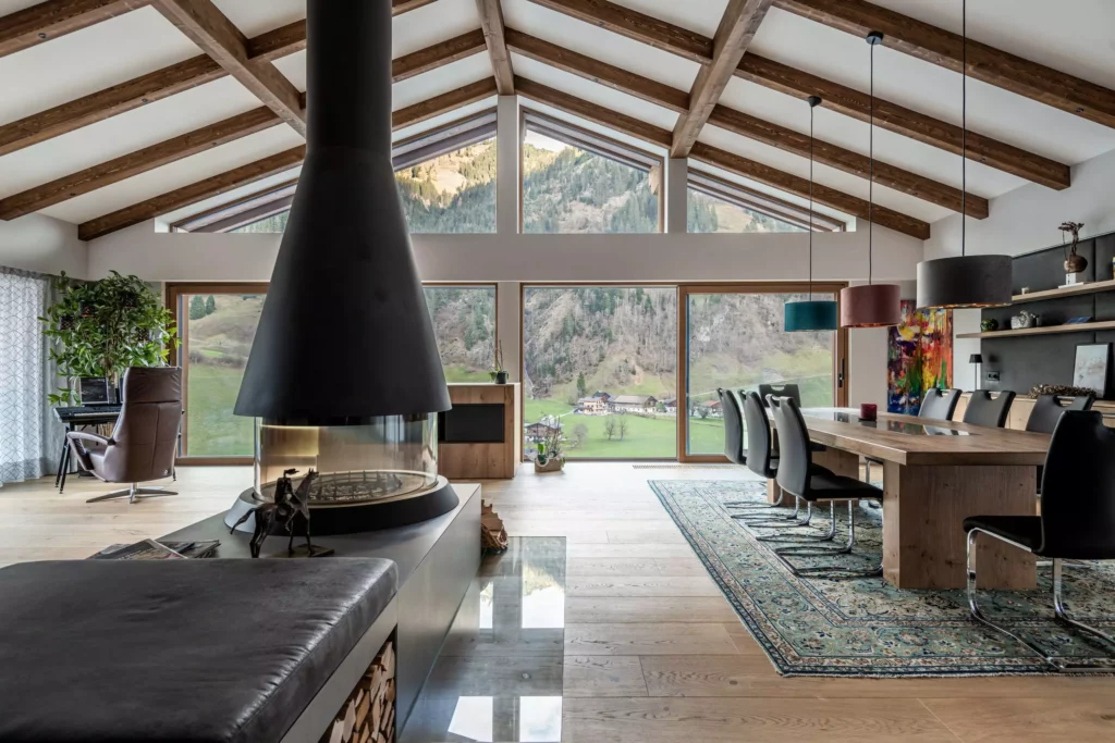 deutinger-projektentwicklung-baumanagement-generalunternehmen-immobilien-salzburg-MFH-Grossarl-Wagnermeyer-17 Moderner Wohn- und Essbereich mit zentralem Kamin, Holzbalkendecke und großflächigen Fenstern mit Bergblick in Grossarl