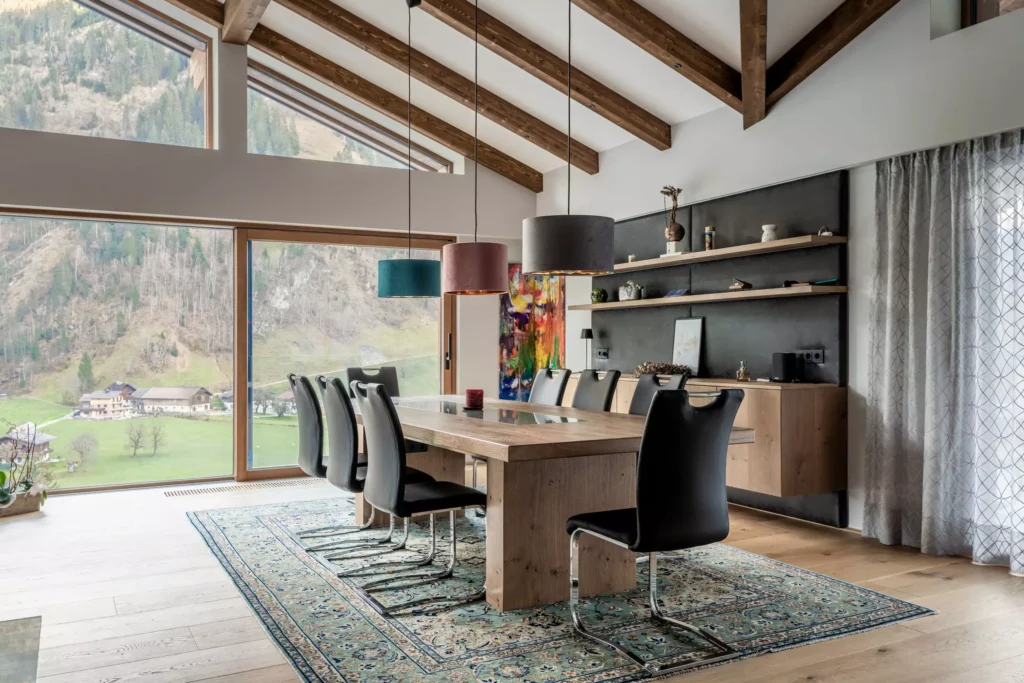 deutinger-projektentwicklung-baumanagement-generalunternehmen-immobilien-salzburg-MFH-Grossarl-Wagnermeyer-18 Moderner Essbereich mit großem Holztisch, schwarzen Stühlen, Teppich und Panoramafenster mit Bergblick in Großarl