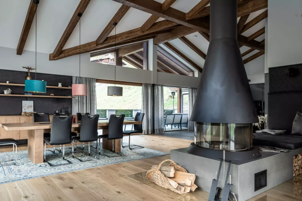 deutinger-projektentwicklung-baumanagement-generalunternehmen-immobilien-salzburg-MFH-Grossarl-Wagnermeyer-20 Moderner Wohn- und Essbereich mit Holzdecke, großem Holztisch, schwarzen Stühlen und zentralem Kamin in Alpenhausstil
