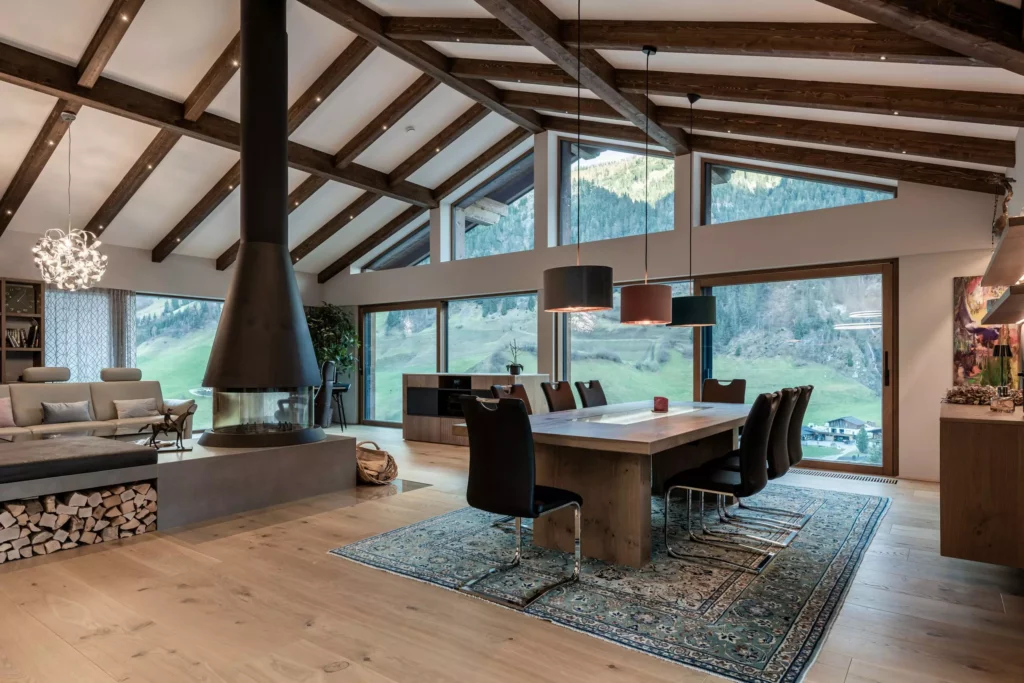 deutinger-projektentwicklung-baumanagement-generalunternehmen-immobilien-salzburg-MFH-Grossarl-Wagnermeyer-22 Moderner Essbereich mit Holztisch, schwarzen Stühlen, Kamin und großen Fenstern mit Bergblick in einem Holzhaus