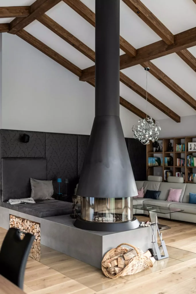 deutinger-projektentwicklung-baumanagement-generalunternehmen-immobilien-salzburg-MFH-Grossarl-Wagnermeyer-24 Moderner Wohnraum mit zentralem Kaminofen, Holzlager, gemütlicher Sitzecke und sichtbaren Holzbalken an der Decke