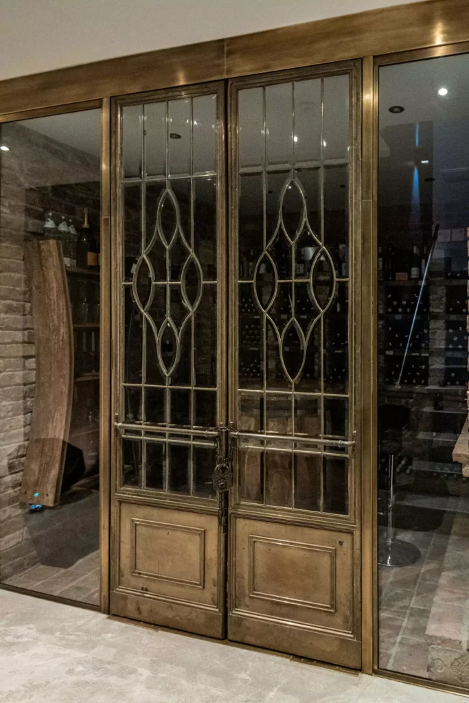deutinger-projektentwicklung-baumanagement-generalunternehmen-immobilien-salzburg-MFH-Grossarl-Wagnermeyer-28 Antike bronzefarbene Doppeltür mit verziertem Glas, dahinter ein Weinkeller mit Weinflaschen und Steinwänden sichtbar.