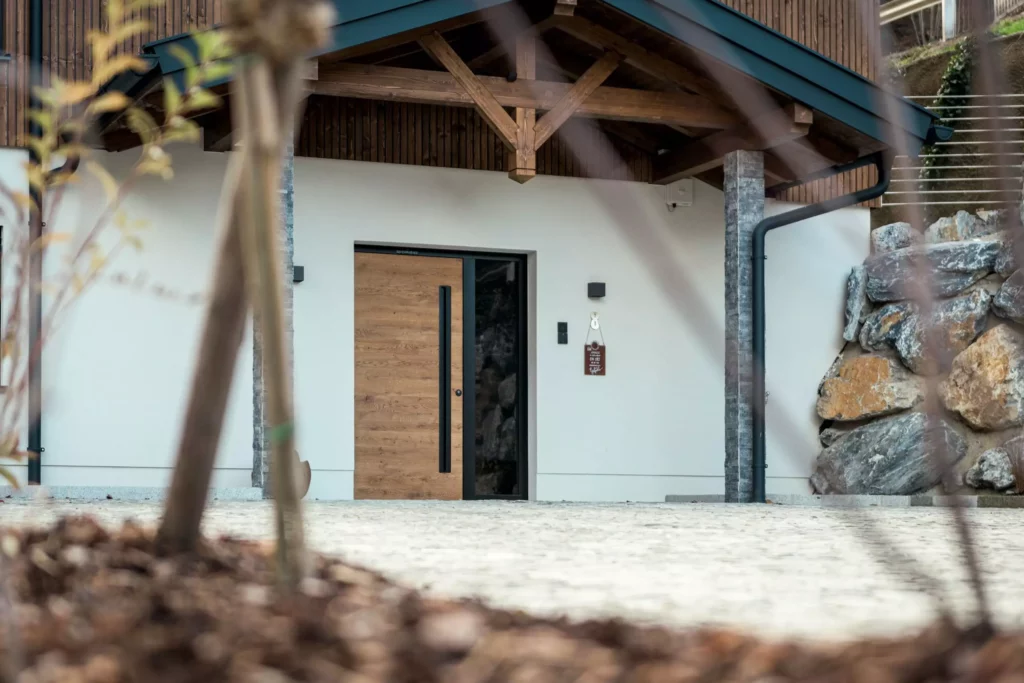 deutinger-projektentwicklung-baumanagement-generalunternehmen-immobilien-salzburg-MFH-Grossarl-Wagnermeyer-9 Eingangsbereich eines modernen Mehrfamilienhauses mit Holzüberdachung, Holztür und Natursteinmauer in Großarl
