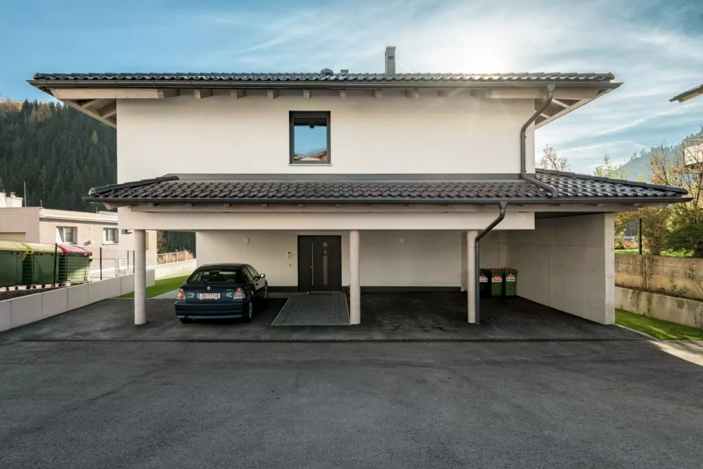 deutinger-projektentwicklung-baumanagement-generalunternehmen-immobilien-salzburg-Mehrfamilienhaus-Altenberger-1 Modernes Mehrfamilienhaus mit Carport, einem geparkten Auto und Mülltonnen, umgeben von Bergen und klarem Himmel in