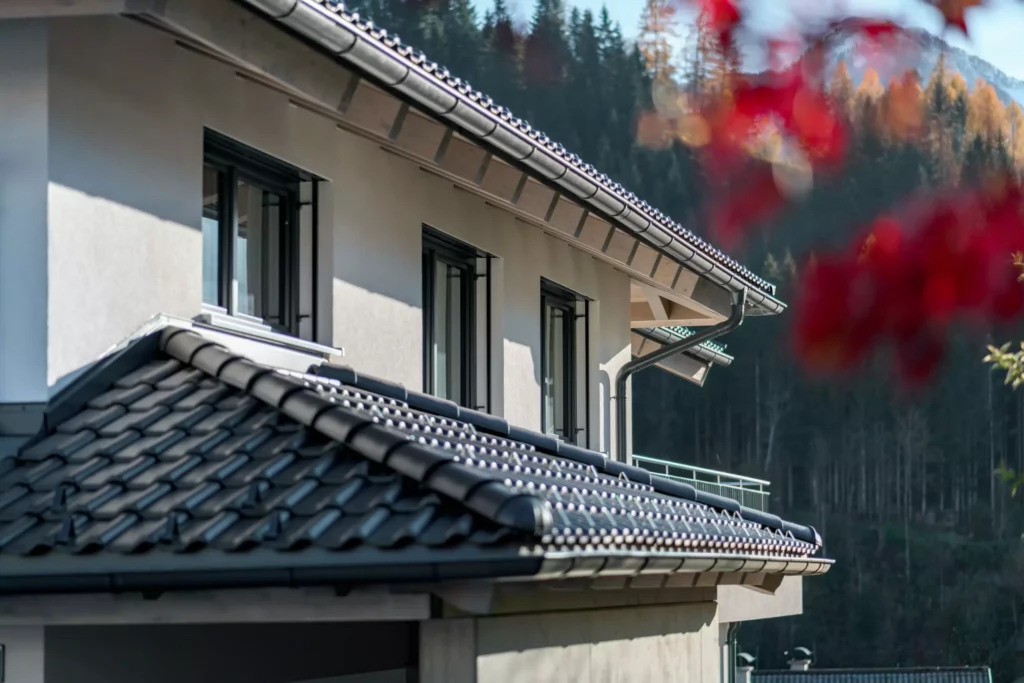 deutinger-projektentwicklung-baumanagement-generalunternehmen-immobilien-salzburg-Mehrfamilienhaus-Altenberger-3 Detailaufnahme eines modernen Mehrfamilienhauses mit grauem Ziegeldach und weißen Wänden vor herbstlichem Wald in Salzburg