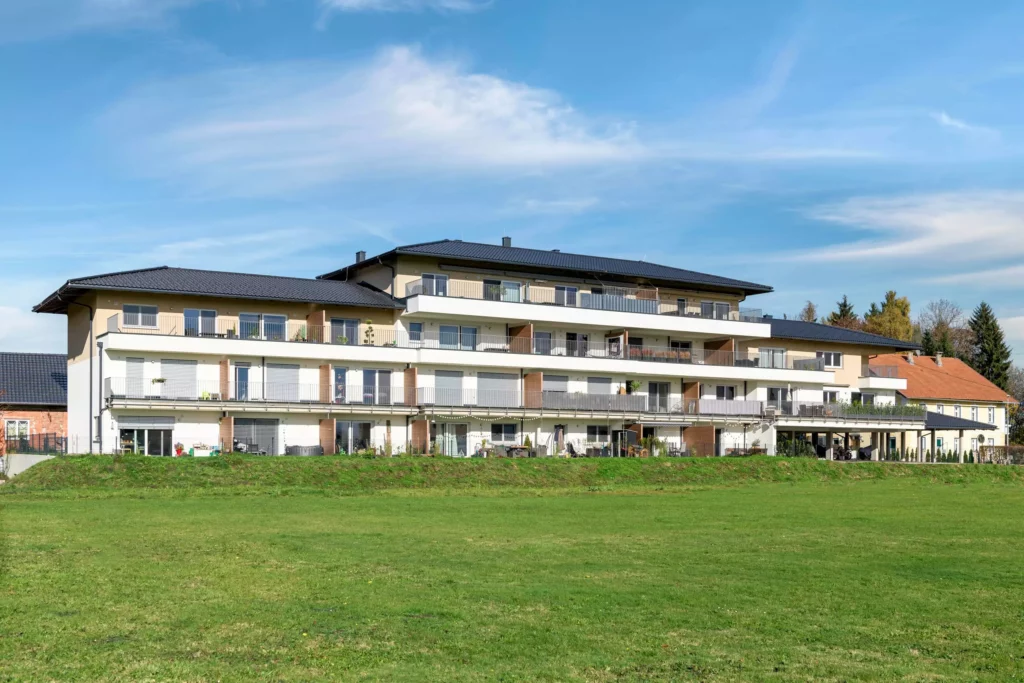 deutinger-projektentwicklung-baumanagement-generalunternehmen-immobilien-salzburg-Panoramapark-Uttendorf-1 Modernes Wohngebäude mit Balkonen vor großem Grünfeld unter blauem Himmel im Panoramapark Uttendorf bei Salzburg