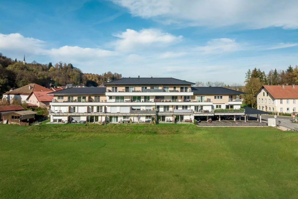 deutinger-projektentwicklung-baumanagement-generalunternehmen-immobilien-salzburg-Panoramapark-Uttendorf-7 Mehrstöckiges Wohngebäude mit Balkonen vor grünem Feld und bewaldetem Hügel unter blauem Himmel im Panoramapark Uttendorf