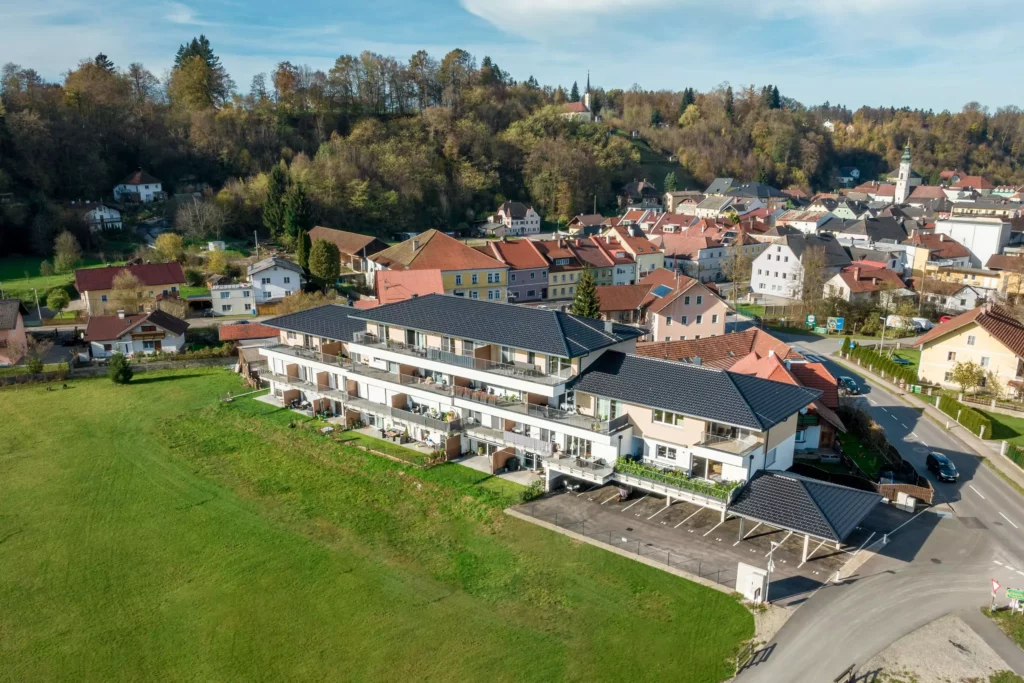 deutinger-projektentwicklung-baumanagement-generalunternehmen-immobilien-salzburg-Panoramapark-Uttendorf-8 Moderner Wohnkomplex mit Balkonen am Ortsrand von Uttendorf vor bewaldetem Hügel und Dorfkern bei klarem Himmel