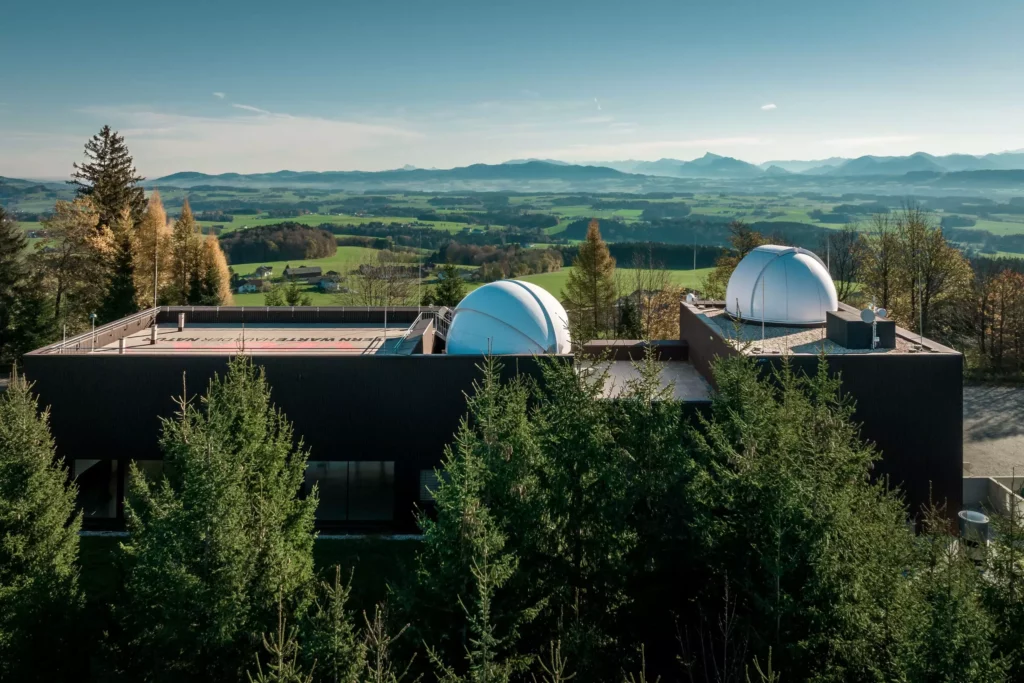 deutinger-projektentwicklung-baumanagement-generalunternehmen-immobilien-salzburg-Sternwarte-Haus-der-Natur-11 Gebäude mit zwei weißen Kuppeln einer Sternwarte umgeben von Bäumen mit Blick auf grüne Landschaft und Berge im Hintergrund