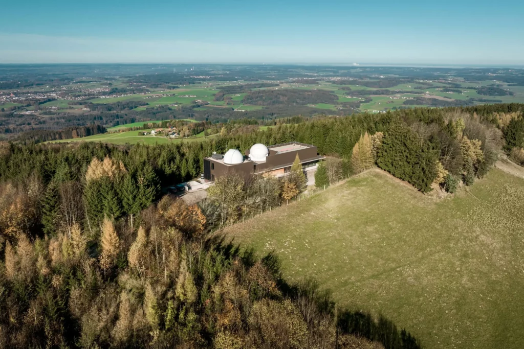 deutinger-projektentwicklung-baumanagement-generalunternehmen-immobilien-salzburg-Sternwarte-Haus-der-Natur-14 Luftaufnahme eines Gebäudes mit zwei Kuppeln in einem Waldgebiet nahe Salzburg, umgeben von Feldern und weiter Landschaft