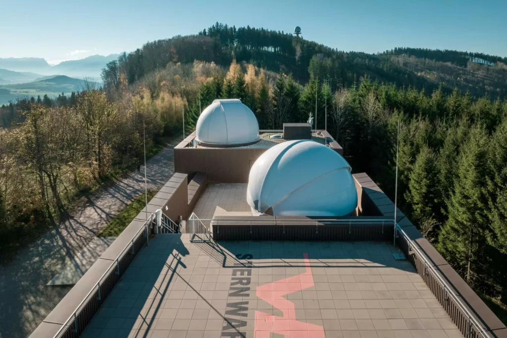 deutinger-projektentwicklung-baumanagement-generalunternehmen-immobilien-salzburg-Sternwarte-Haus-der-Natur-15 Dachterrasse mit zwei weißen Kuppeln einer Sternwarte umgeben von Wald und Bergen im Hintergrund bei klarem Himmel