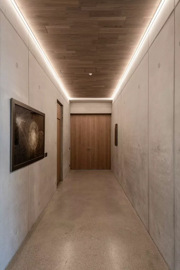 deutinger-projektentwicklung-baumanagement-generalunternehmen-immobilien-salzburg-Sternwarte-Haus-der-Natur-16 Moderner Flur mit Betonwänden, Holzdecke, indirekter Beleuchtung und zwei Holztüren am Ende des Gangs