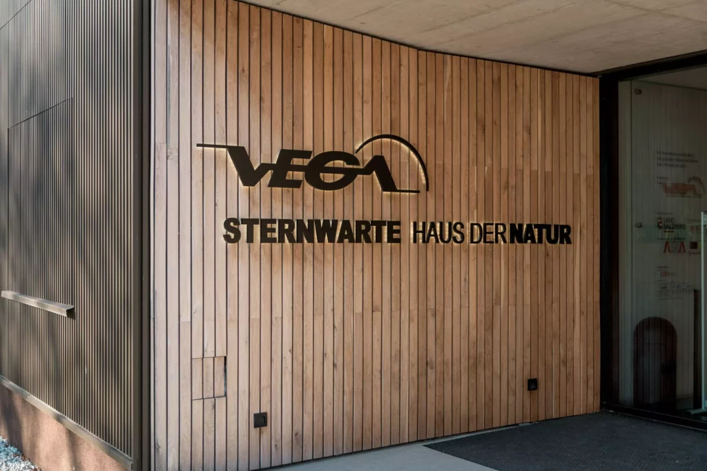 deutinger-projektentwicklung-baumanagement-generalunternehmen-immobilien-salzburg-Sternwarte-Haus-der-Natur-2 Eingang mit Holzverkleidung und schwarzem Schriftzug VEGA Sternwarte Haus der Natur in Salzburg, modernes