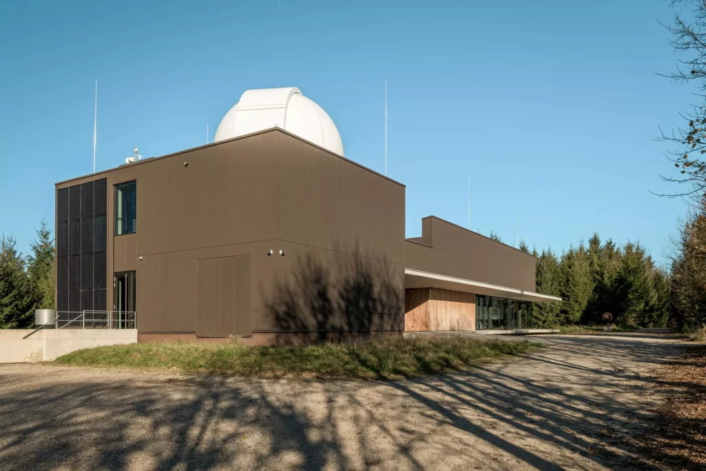 deutinger-projektentwicklung-baumanagement-generalunternehmen-immobilien-salzburg-Sternwarte-Haus-der-Natur-3 Modernes Bauprojekt mit braunem Gebäude und weißer Sternwartekuppel umgeben von Wald bei klarem Himmel