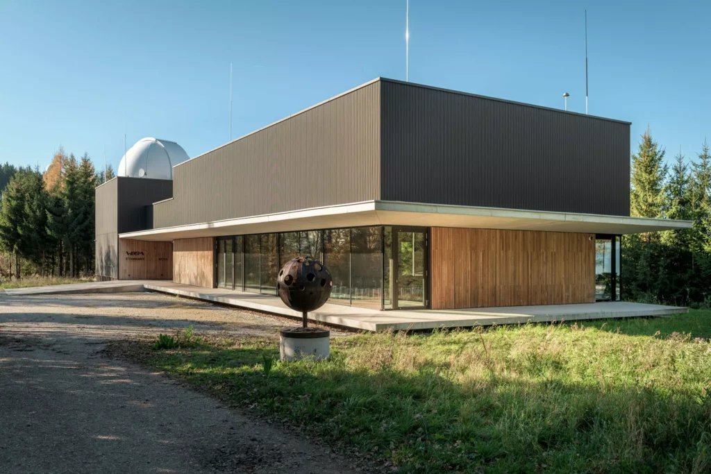 deutinger-projektentwicklung-baumanagement-generalunternehmen-immobilien-salzburg-Sternwarte-Haus-der-Natur-5 Modernes Gebäude mit Holz- und Glasfassade, Sternwarte mit Kuppel im Hintergrund, umgeben von Bäumen und Wiese