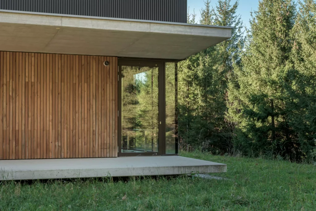 deutinger-projektentwicklung-baumanagement-generalunternehmen-immobilien-salzburg-Sternwarte-Haus-der-Natur-6 Moderner Gebäudeeingang mit Holzverkleidung, Glasfront und Betonpodest vor Waldkulisse in Salzburg