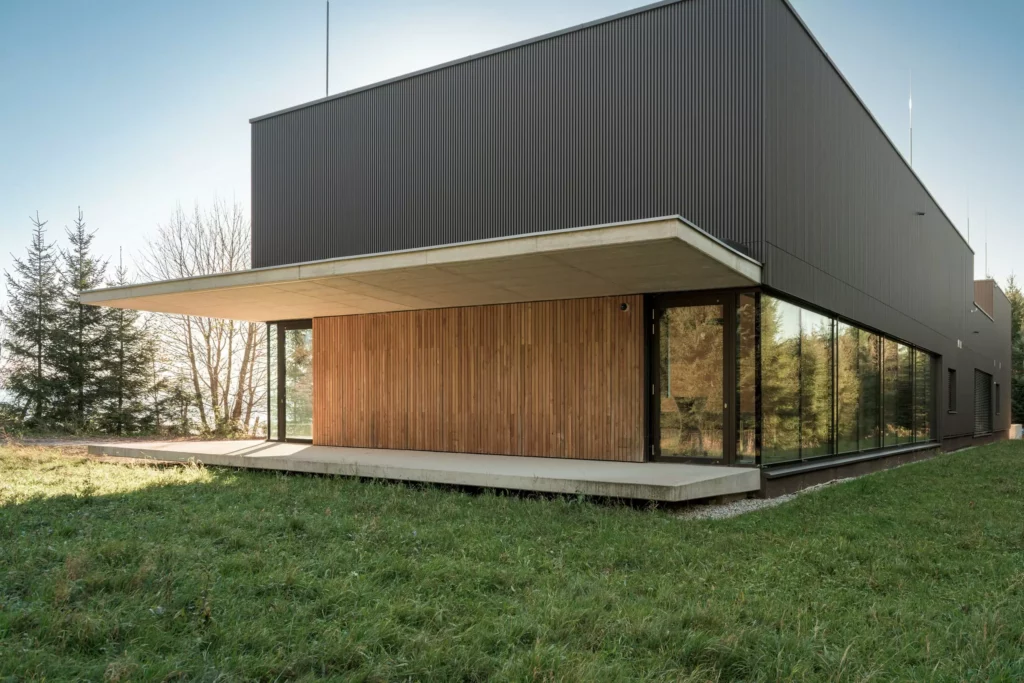 deutinger-projektentwicklung-baumanagement-generalunternehmen-immobilien-salzburg-Sternwarte-Haus-der-Natur-7 Modernes Gebäude mit Holz- und Glasfassade umgeben von Wiese und Bäumen bei klarem Himmel in Salzburg