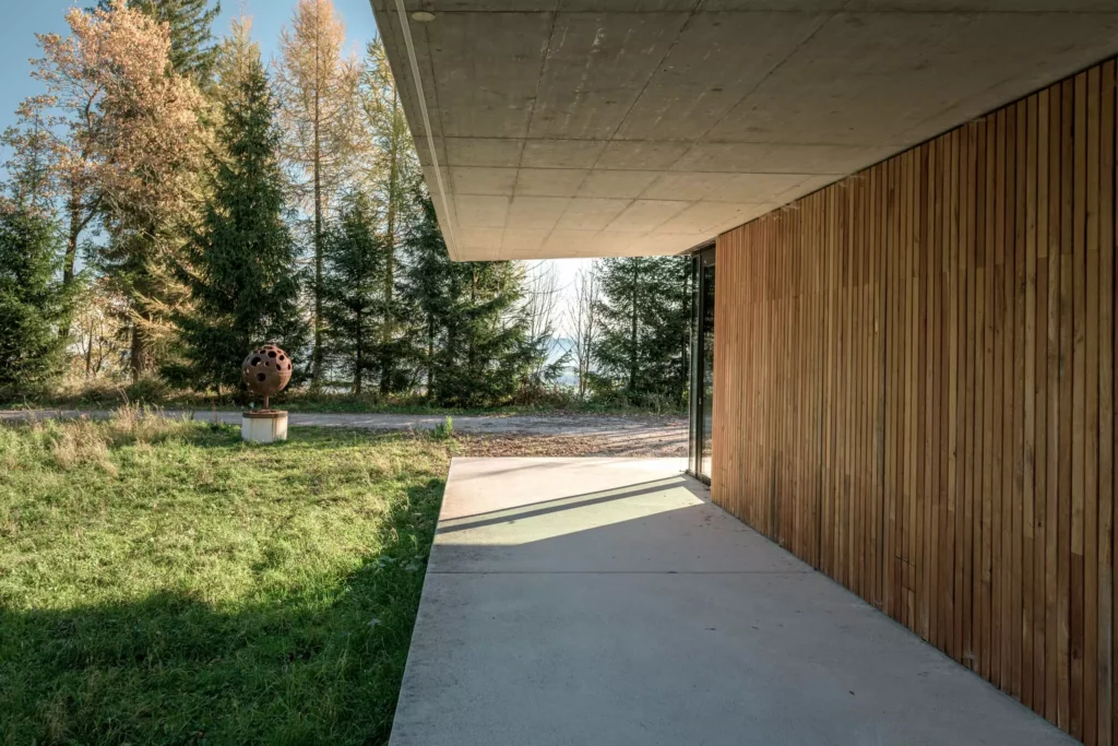 deutinger-projektentwicklung-baumanagement-generalunternehmen-immobilien-salzburg-Sternwarte-Haus-der-Natur-8 Moderner Gebäudeeingang mit Holzverkleidung und Betonüberhang, umgeben von Wiese und Nadelbäumen bei Sonnenschein