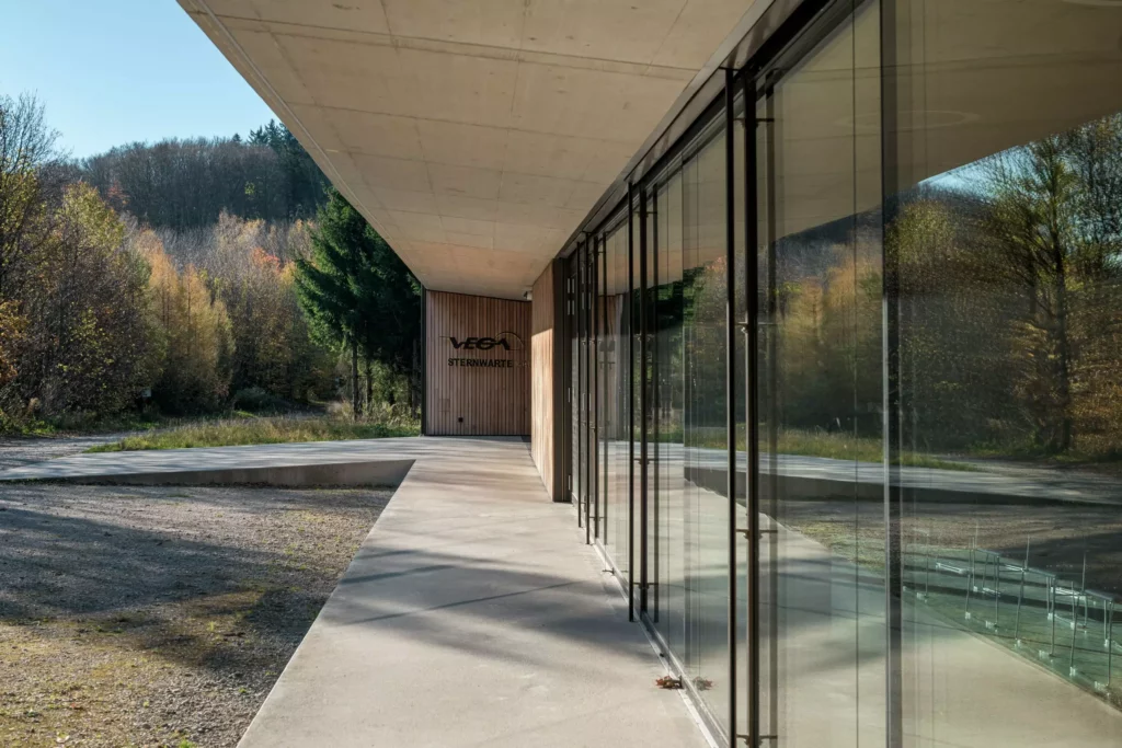 deutinger-projektentwicklung-baumanagement-generalunternehmen-immobilien-salzburg-Sternwarte-Haus-der-Natur-9 Moderner Gebäudeeingang mit großer Glasfassade und Holzverkleidung, umgeben von herbstlichem Wald in Salzburg