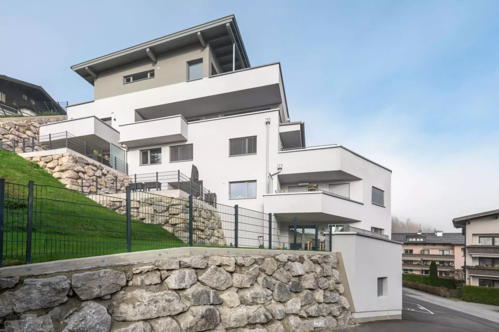 deutinger-projektentwicklung-baumanagement-generalunternehmen-immobilien-salzburg-Wohnpark-Bischofshofen-1 Moderner Wohnpark mit weißen Mehrfamilienhäusern und Natursteinmauern in Bischofshofen, Salzburg bei klarem Himmel