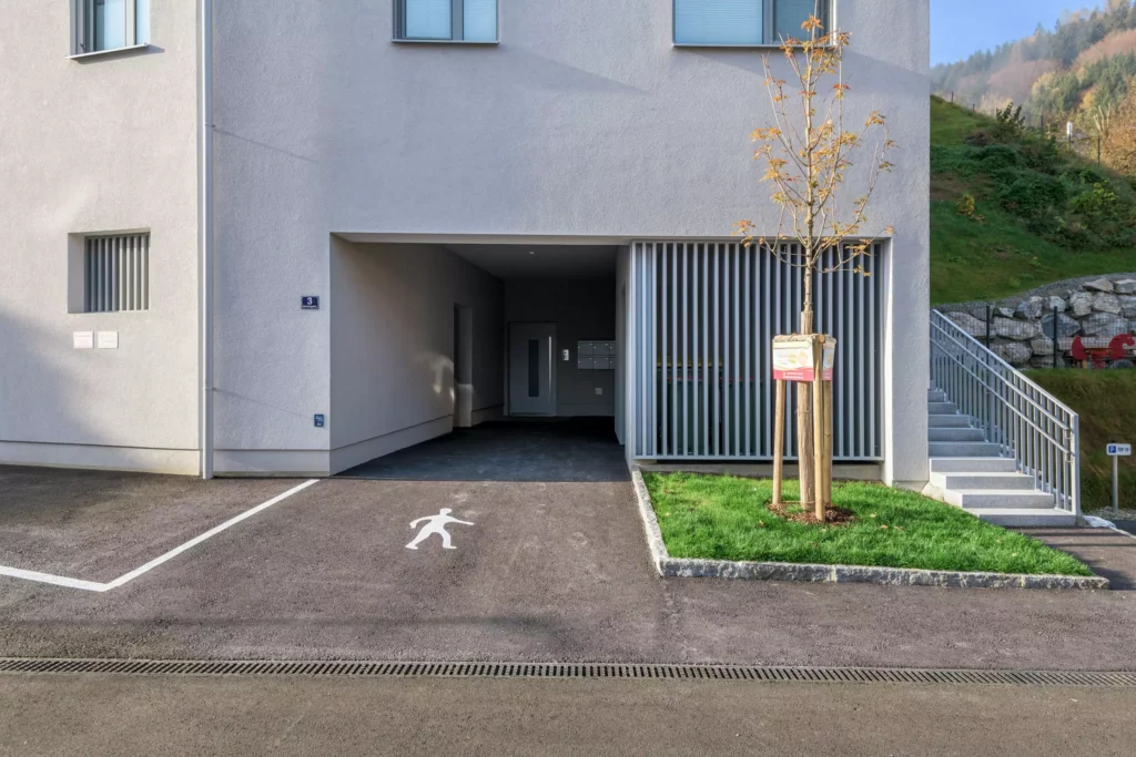 deutinger-projektentwicklung-baumanagement-generalunternehmen-immobilien-salzburg-Wohnpark-Bischofshofen-3 Eingang zu einem modernen Wohngebäude mit Parkfläche, Baum und Treppe im Wohnpark Bischofshofen, Salzburg.
