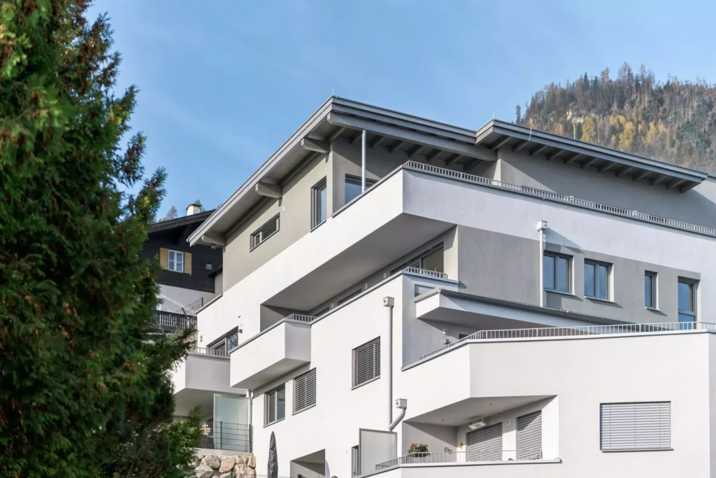 deutinger-projektentwicklung-baumanagement-generalunternehmen-immobilien-salzburg-Wohnpark-Bischofshofen-4 Moderner Wohnpark mit weißen und grauen Fassaden, Balkonen und Fensterläden vor bewaldetem Berg in Bischofshofen bei Salzburg