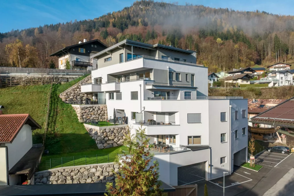 deutinger-projektentwicklung-baumanagement-generalunternehmen-immobilien-salzburg-Wohnpark-Bischofshofen-5 Moderner Wohnpark mit weißen Mehrfamilienhäusern und Terrassen in Bischofshofen vor bewaldetem Berghang im Herbst