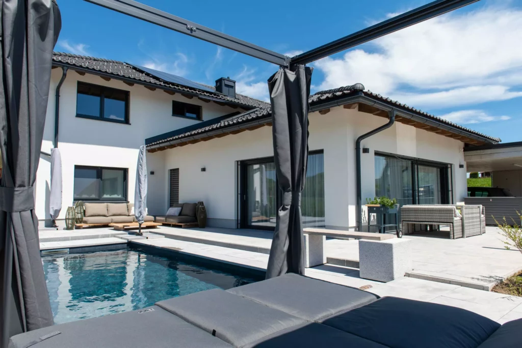 deutinger-projektentwicklung-baumanagement-generalunternehmen-immobilien-salzburg-projekt-einfamilienhaus-31 Modernes Einfamilienhaus mit Pool, Terrasse und Lounge-Möbeln bei sonnigem Himmel, Beispiel für Bauprojektmanagement und