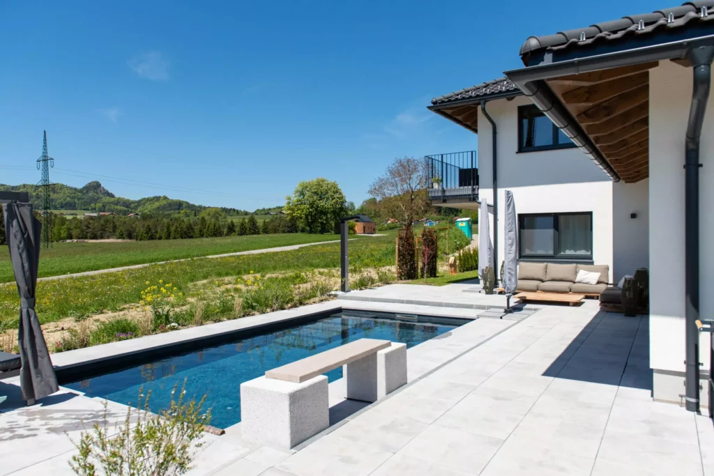 deutinger-projektentwicklung-baumanagement-generalunternehmen-immobilien-salzburg-projekt-einfamilienhaus-34 Moderner Außenbereich eines Einfamilienhauses mit Pool, Sitzbank und Sofa vor ländlicher Kulisse bei klarem Himmel