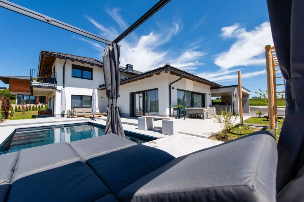 deutinger-projektentwicklung-baumanagement-generalunternehmen-immobilien-salzburg-projekt-einfamilienhaus-35 Modernes Einfamilienhaus mit Pool und Terrasse unter blauem Himmel, Sicht von einer überdachten Lounge aus