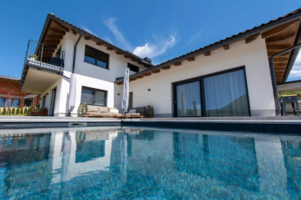 deutinger-projektentwicklung-baumanagement-generalunternehmen-immobilien-salzburg-projekt-einfamilienhaus-36 Modernes Einfamilienhaus mit Terrasse, Lounge-Möbeln und klarem Pool unter blauem Himmel in Salzburg