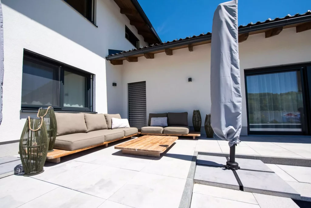 deutinger-projektentwicklung-baumanagement-generalunternehmen-immobilien-salzburg-projekt-einfamilienhaus-37 Moderne Terrasse mit grauem Sofa, Holztisch, zwei Laternen und geschlossenem Sonnenschirm vor weißem Einfamilienhaus