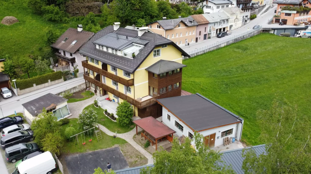 deutinger-projektentwicklung-baumanagement-generalunternehmen-immobilien-salzburg-wohnanlage-werfen64-02 Mehrstöckiges Wohngebäude mit gelber Fassade und angrenzendem Flachdachbau in grüner Umgebung in Werfen, Salzburg