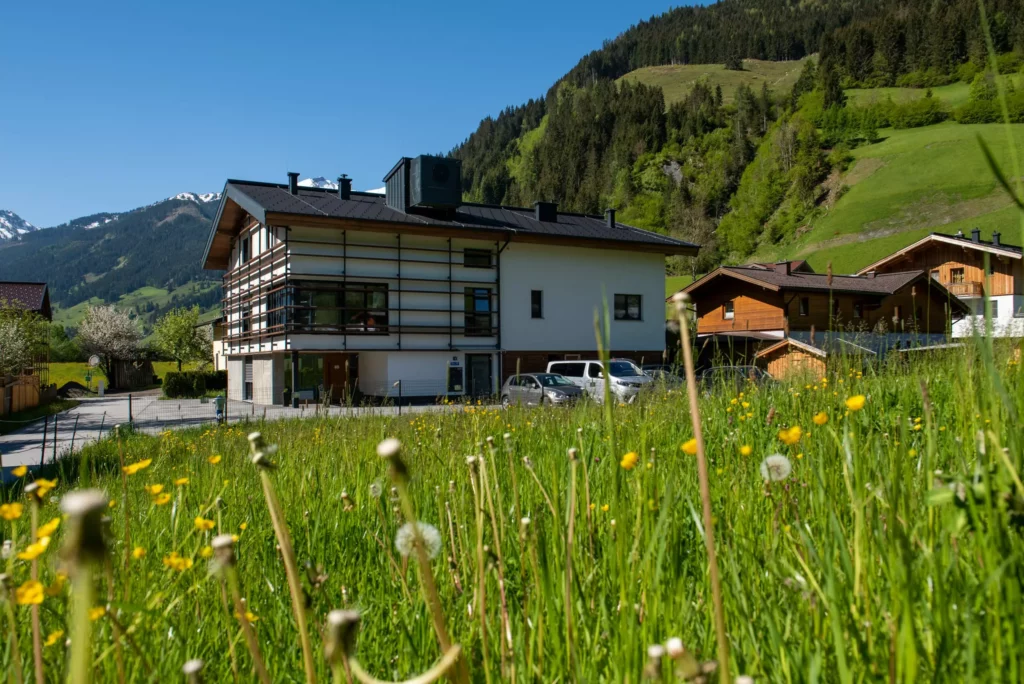 deutinger-projektentwicklung-baumanagement-generalunternehmen-immobilien-salzburg-wohnhaus-rohrmoser-05 Modernes Wohnhaus mit großem Fensterfront in grüner Berglandschaft bei Rohrmoser, Salzburg, unter blauem Himmel