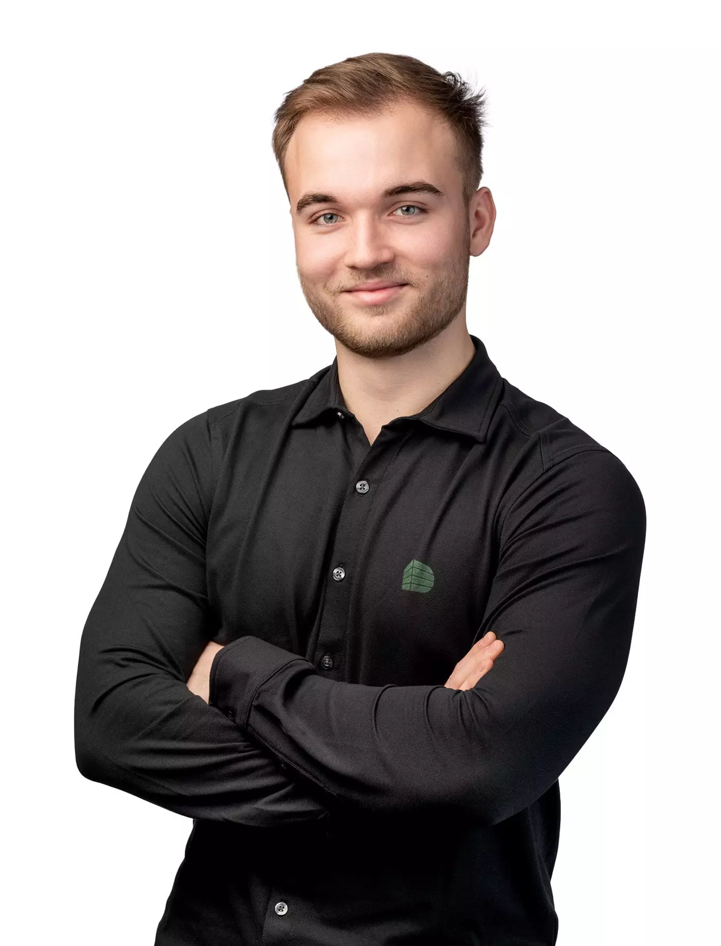 Junger Mann mit kurzem Haar und Bart, trägt schwarzes Hemd mit grünem Logo, Arme verschränkt, vor weißem Hintergrund.