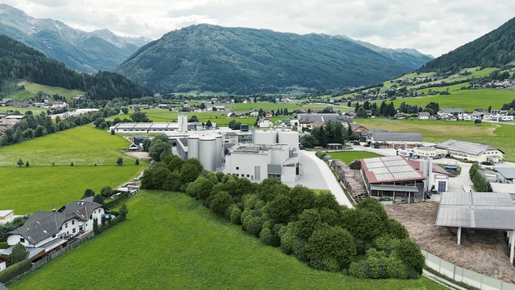 Luftaufnahme eines Industriegebiets mit modernen Gebäuden, umgeben von grünen Wiesen und Bergen im Hintergrund