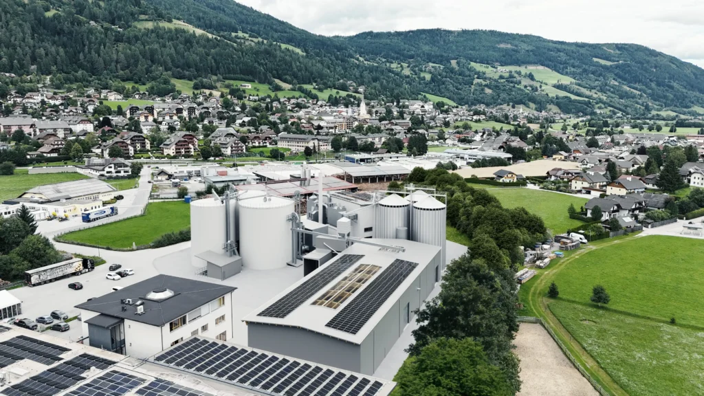 02_RZ Pellets Luftaufnahme eines Industriegebäudes mit Solaranlagen auf dem Dach, umgeben von grünen Feldern und einem Dorf vor bewaldeten