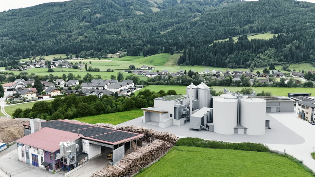 03_RZ Pellets Industrieanlage zur Pelletproduktion mit Holzstapeln im Vordergrund und Dorf vor bewaldetem Bergpanorama im Hintergrund