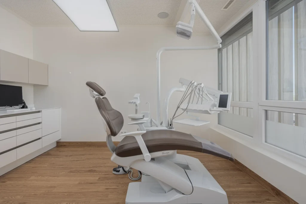 260526_Deutinger-DrNachtigal-©sobietzki-low-11 Moderner Behandlungsstuhl in heller, minimalistisch eingerichteter Zahnarztpraxis mit Holzboden und großen Fenstern.