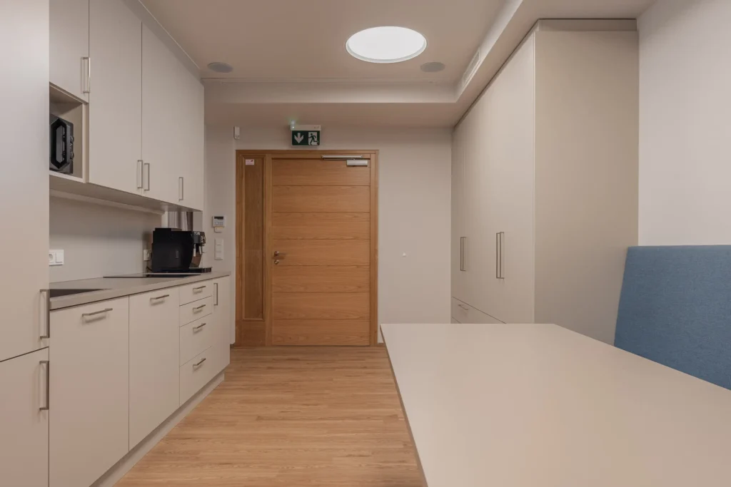 260526_Deutinger-DrNachtigal-©sobietzki-low-19 Moderne Küchenzeile mit weißen Schränken, Kaffeemaschine, Holztür und blau gepolsterter Sitzbank in hellem Raum mit