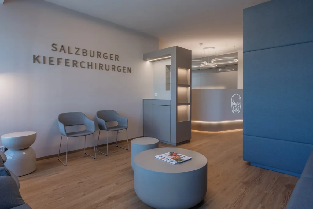 260526_Deutinger-DrNachtigal-©sobietzki-low-6 Moderne Wartezone mit grauen Stühlen, runden Tischen und Schriftzug Salzburger Kieferchirurgen an der Wand